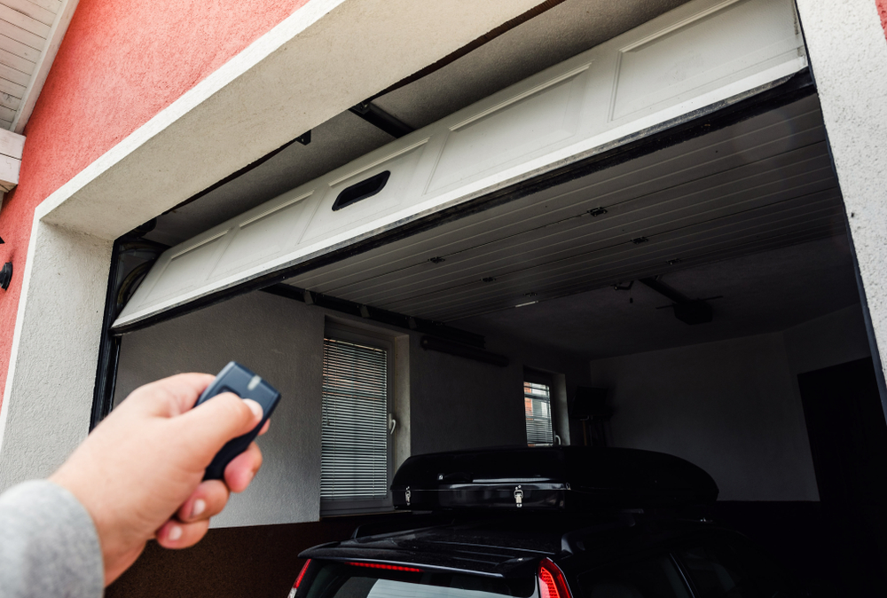 A hand holds a garage door remote, pointing at a partially open garage door.